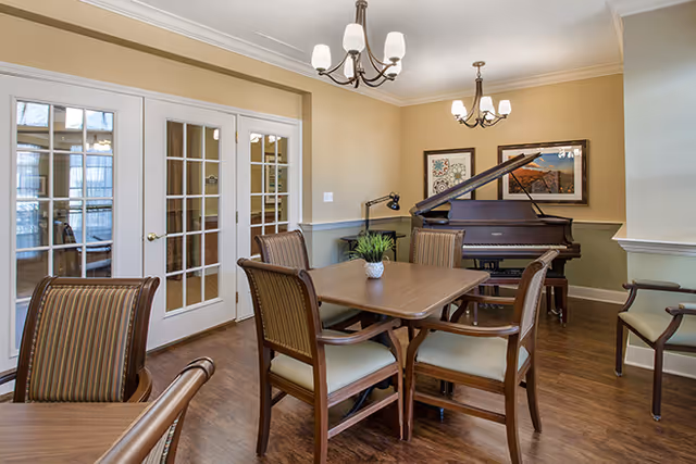 A well-lit room with wooden flooring featuring a square wooden table surrounded by six cushioned chairs. In the background, there is a grand piano with a framed picture above it, and two chandeliers hanging from the ceiling. French doors with glass panes are on the left side of the room.