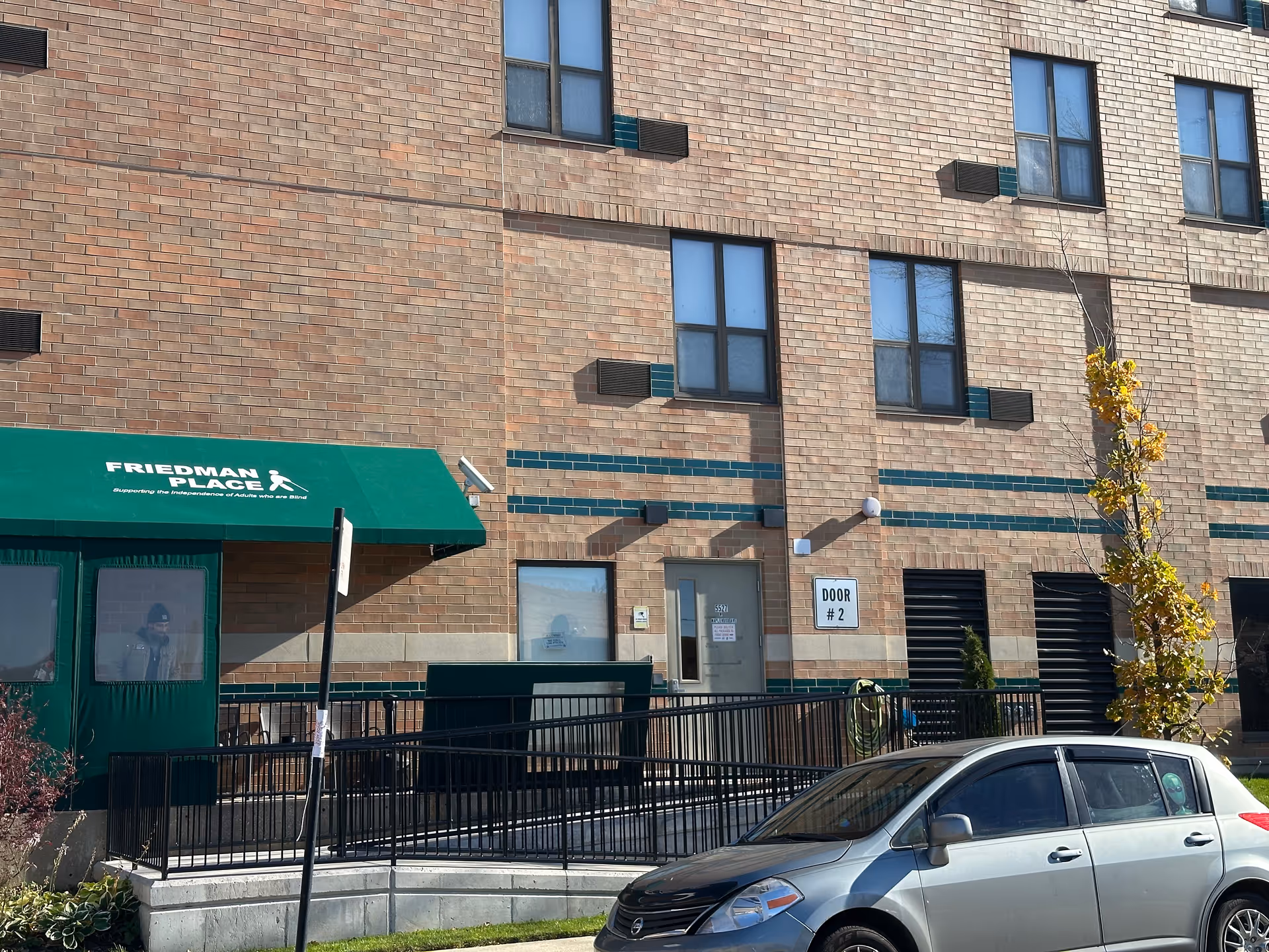Exterior view of a brick building with a green awning labeled 'Friedman Place supporting the independence of adults who are blind.' There is a ramp with black railings leading to a door marked 'Door #2.' A silver car is parked in front of the building, and a small tree with yellow leaves is visible on the right side.