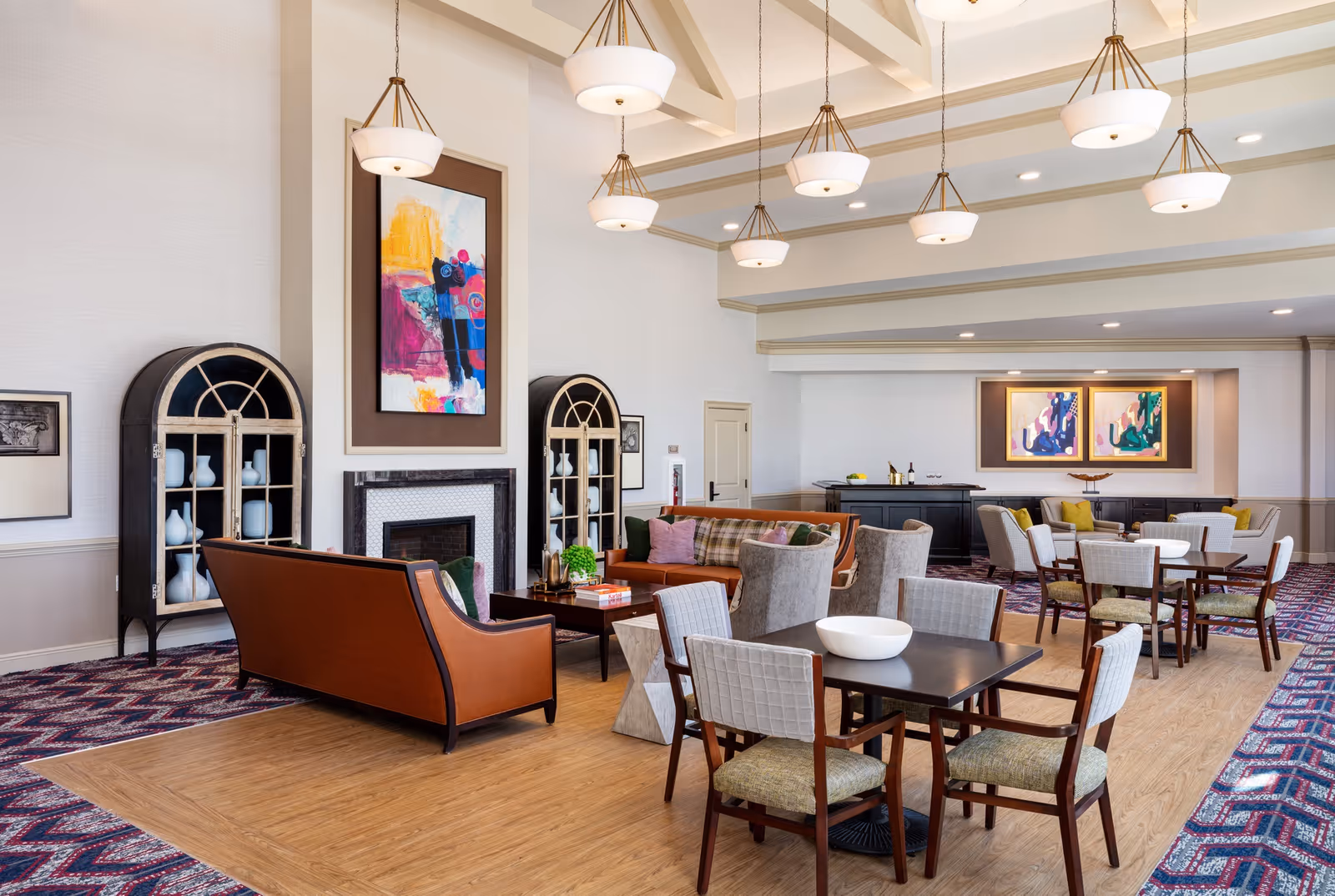 A spacious and well-lit common area in a senior living facility featuring multiple seating arrangements including brown leather sofas, cushioned chairs, and tables with white bowls. The room has a high ceiling with multiple pendant lights, colorful abstract paintings on the walls, and decorative cabinets with white vases. The floor combines wood and patterned carpet.