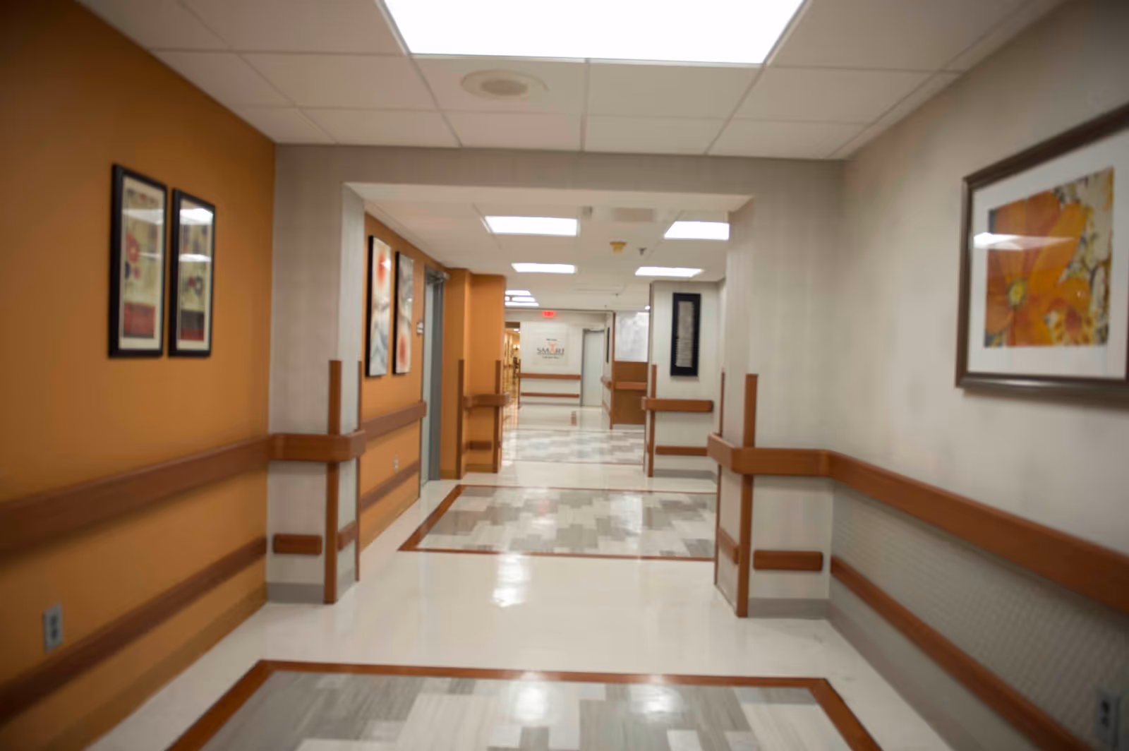 A clean and well-lit hallway in a healthcare facility with handrails on both sides, framed artwork on the walls, and a tiled floor with a patterned design. The hallway leads to another corridor with more artwork and doors visible in the distance.
