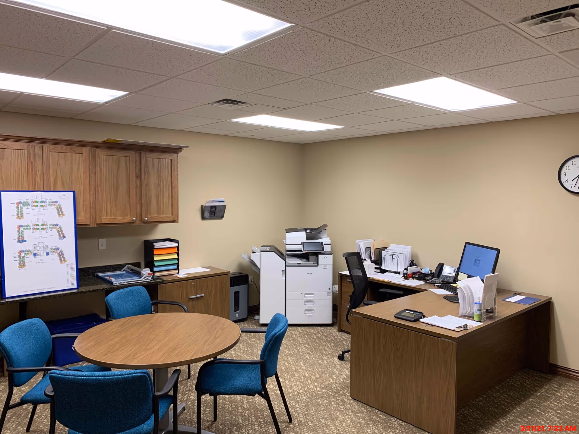 An interior office with a round meeting table and blue chairs, desks with a computer and paperwork, and a large copier against beige walls.