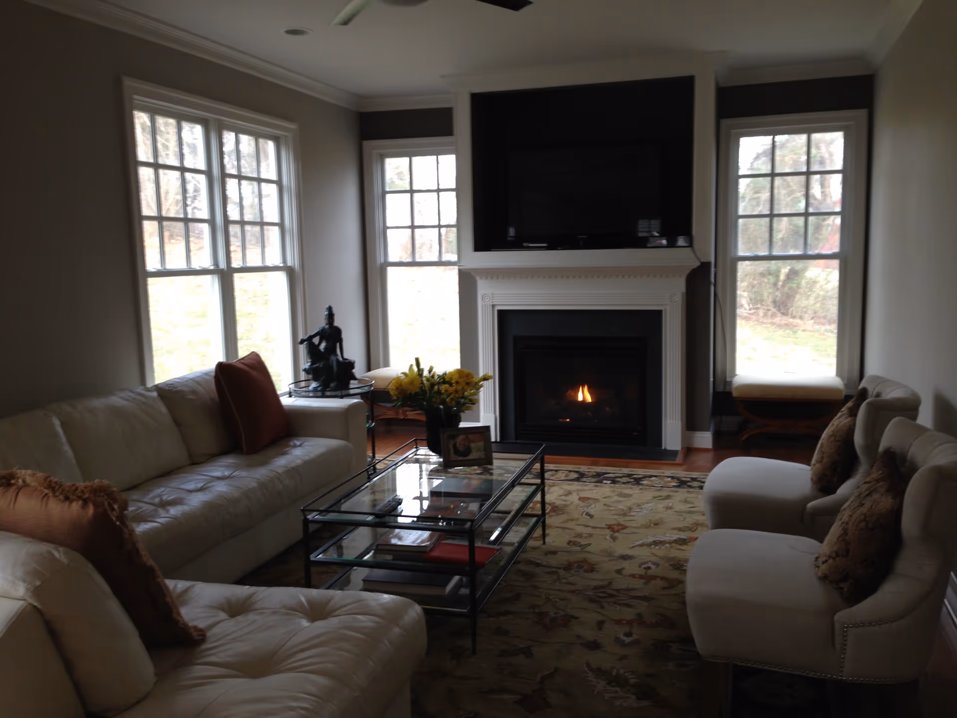 A cozy living room with a lit fireplace beneath a mounted flat-screen TV. The room features two large windows on either side of the fireplace, a white leather sofa with brown pillows on the left, two beige upholstered chairs with patterned cushions on the right, a glass coffee table with books and a flower vase, and a decorative statue on a side table near the window.
