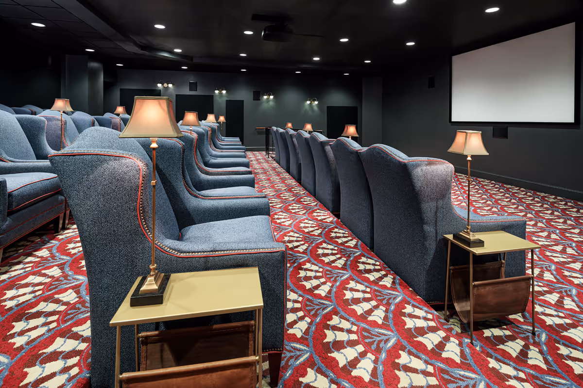 Interior view of a theater room with rows of blue upholstered armchairs, small side tables with lamps, and a large projection screen at the front. The room has a patterned red, white, and blue carpet and dim lighting.