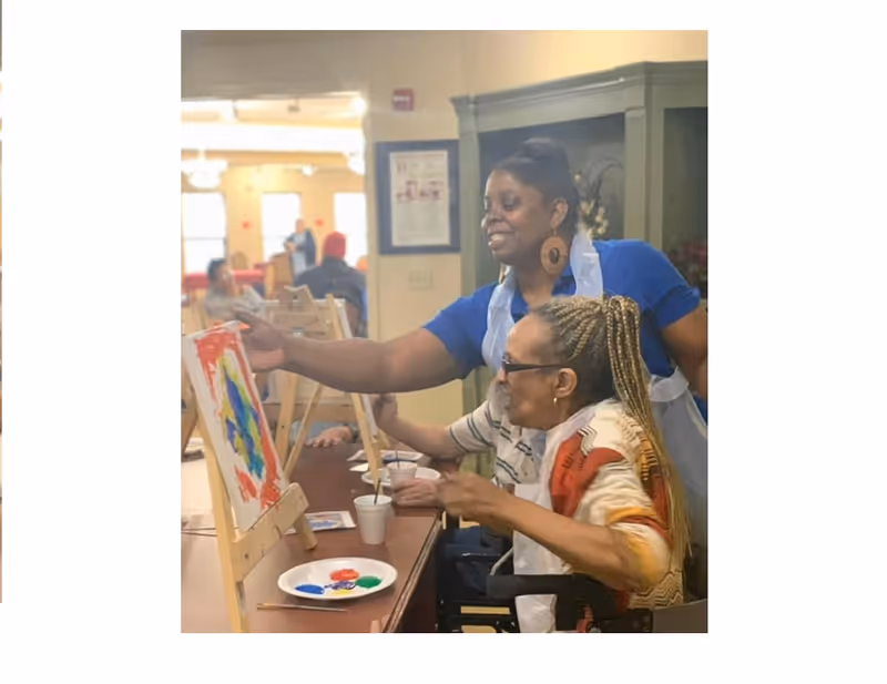 Two women engaged in a painting activity at a table indoors. One woman, standing and wearing a blue shirt and apron, is assisting the other woman, who is seated and wearing glasses and a colorful sweater, with her painting on an easel. There are paint cups and a palette with paint on the table. Other people and tables are visible in the background in a well-lit room.
