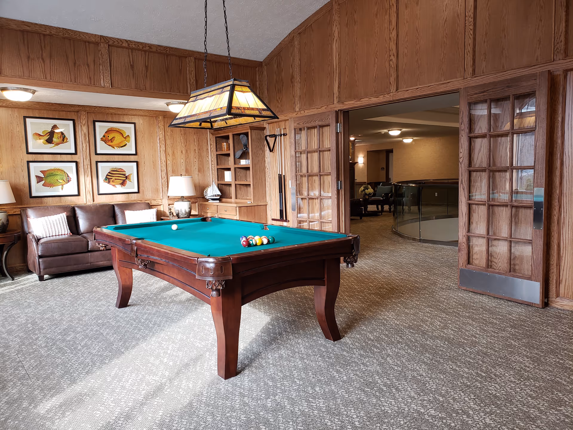 A cozy indoor recreational room with a green felt pool table in the center, surrounded by wood-paneled walls. There is a brown leather couch with two pillows against the wall, four framed fish illustrations above it, and a hanging stained glass light fixture above the pool table. The room has carpeted flooring and an open doorway with glass-paneled wooden doors leading to another seating area.