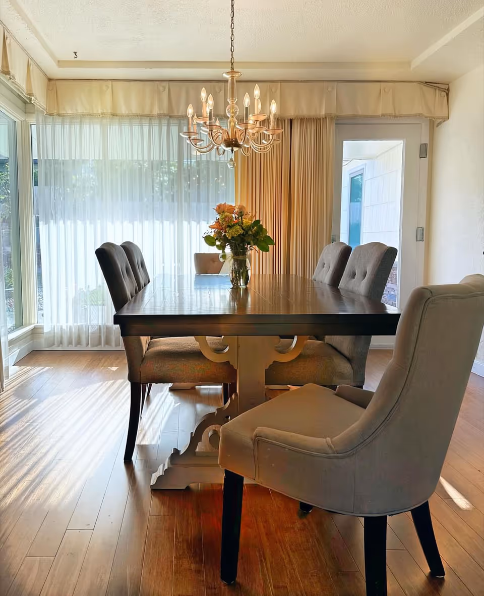 A bright dining room with a wooden dining table surrounded by six upholstered chairs. A vase with flowers is placed in the center of the table. A chandelier with lit bulbs hangs above the table. Large windows with sheer curtains allow natural light to fill the room, and there is a door with a window on the right side.