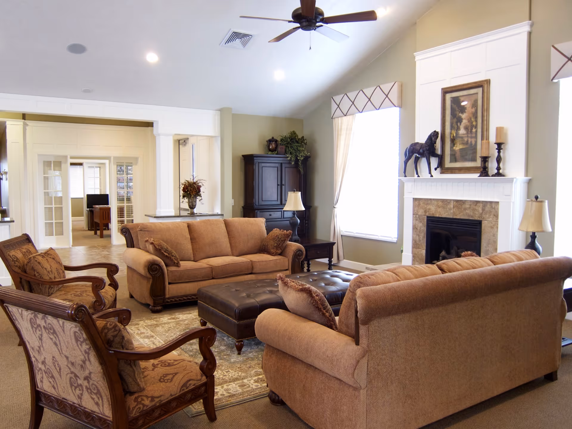 A cozy living room with two beige sofas, two patterned armchairs, a large tufted leather ottoman, and a fireplace with decorative items including a horse statue and candles. The room has large windows with curtains, a ceiling fan, and a cabinet with plants and a lamp.