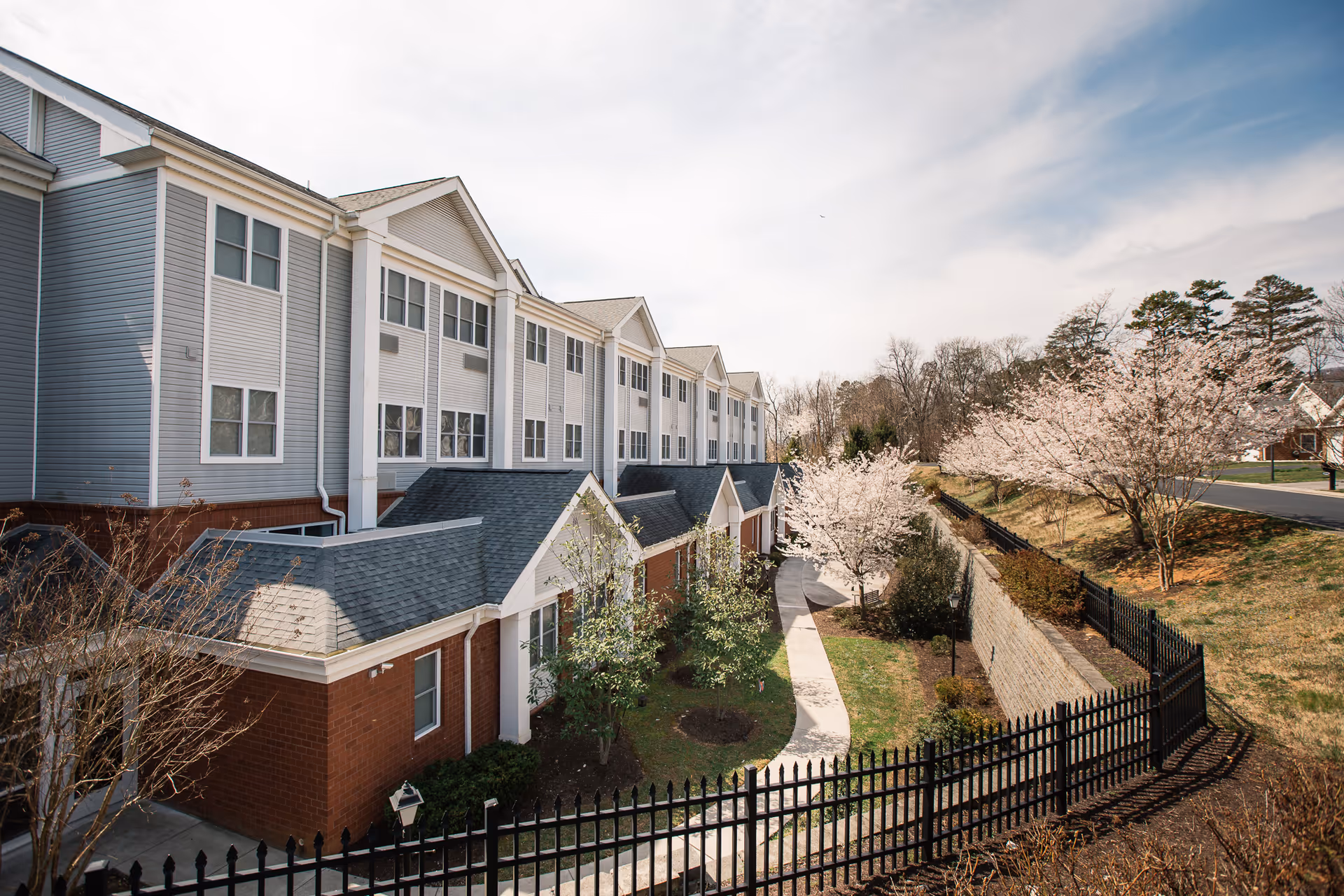 Exterior view of a multi-story senior living facility building with a brick and siding facade, a paved walkway, landscaped greenery, and blooming trees along a black metal fence under a partly cloudy sky.