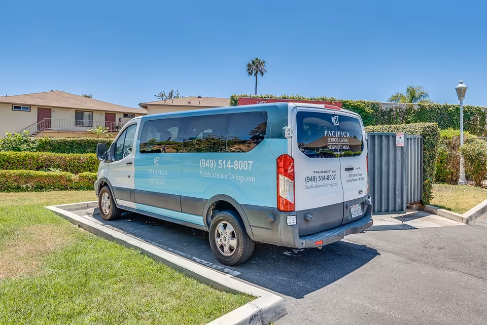 A blue and white shuttle van parked in a parking lot near a grassy area and residential buildings. The van has signage for Pacifica Senior Living Newport Mesa with a phone number and website on its side and rear windows.