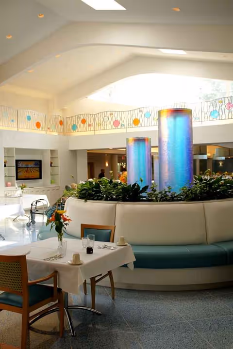 Bright dining area with white tablecloths, chairs, and a curved planter centerpiece in a modern atrium.