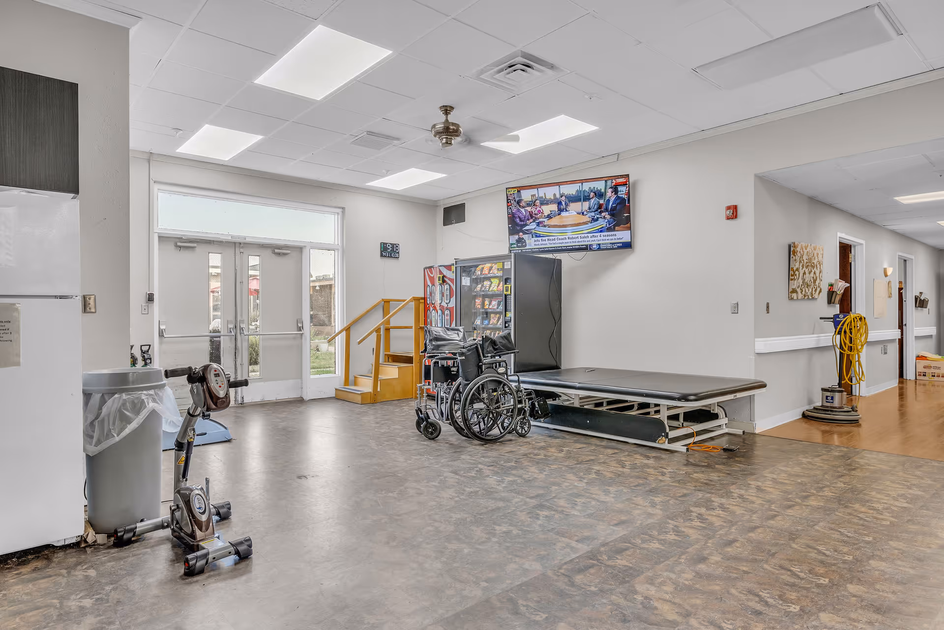 Interior view of a rehabilitation center room with exercise equipment including a stationary bike, a wheelchair, a therapy table, a vending machine, and a wall-mounted television. There is a double glass door leading outside, and a hallway with cleaning equipment is visible to the right.