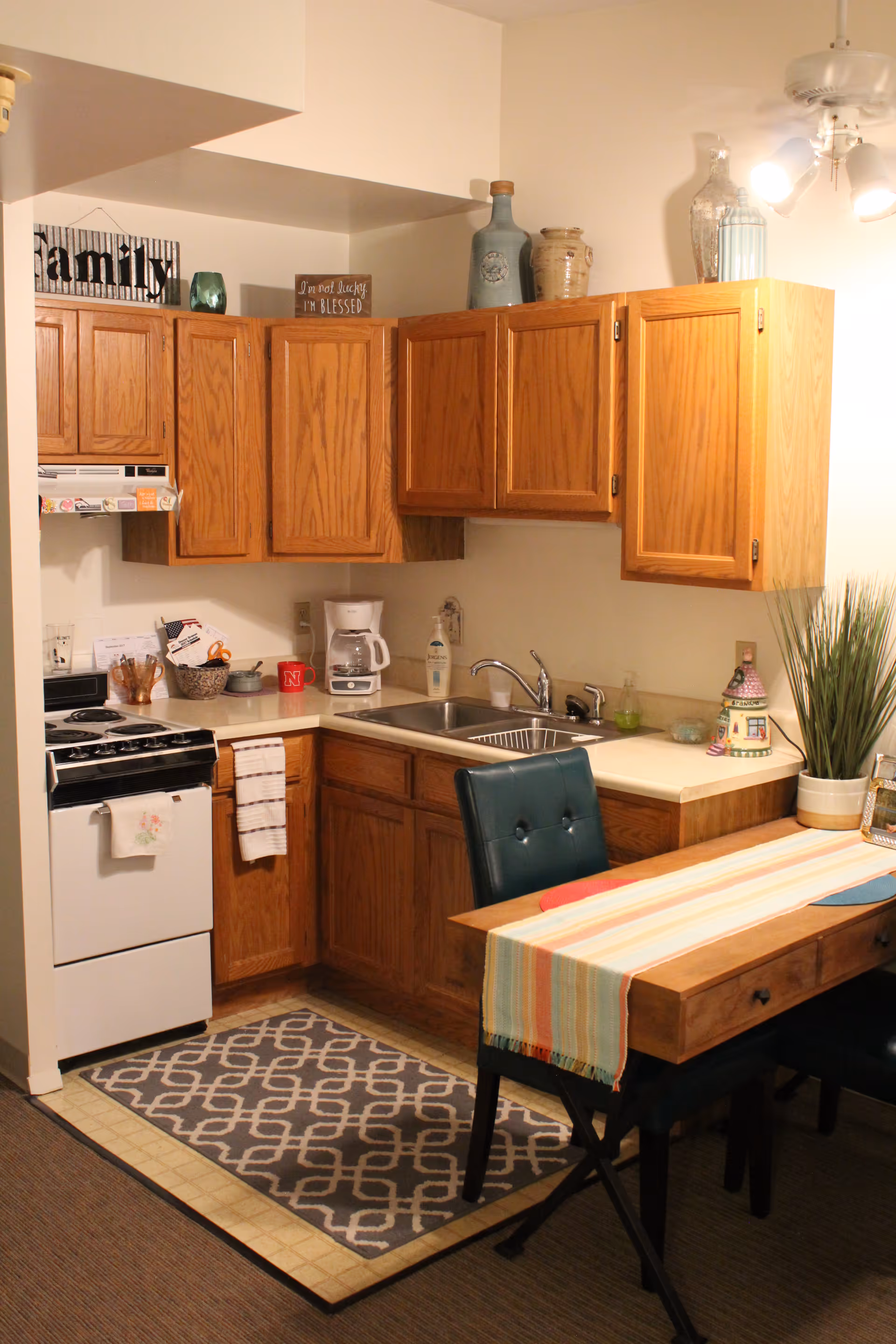 A small kitchen area with wooden cabinets, a white stove, a coffee maker, a double sink, and a dining table with a colorful striped table runner and dark green chairs. Decorative items are placed on top of the cabinets and on the counter, including a plant and a small house-shaped ornament.