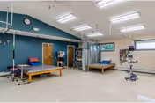 A spacious physical therapy or rehabilitation room with two therapy tables, exercise equipment, and various therapy tools hanging on the walls. The room has a high ceiling with fluorescent lights, a clock on the wall, and a window letting in natural light.