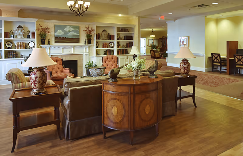 A cozy living room area in a senior living facility with leather sofas, wooden side tables with decorative lamps, a wooden console table with flower vases and bird figurines, and built-in white bookshelves surrounding a fireplace. The room has warm lighting and a wooden floor, with a glimpse of a dining area in the background.