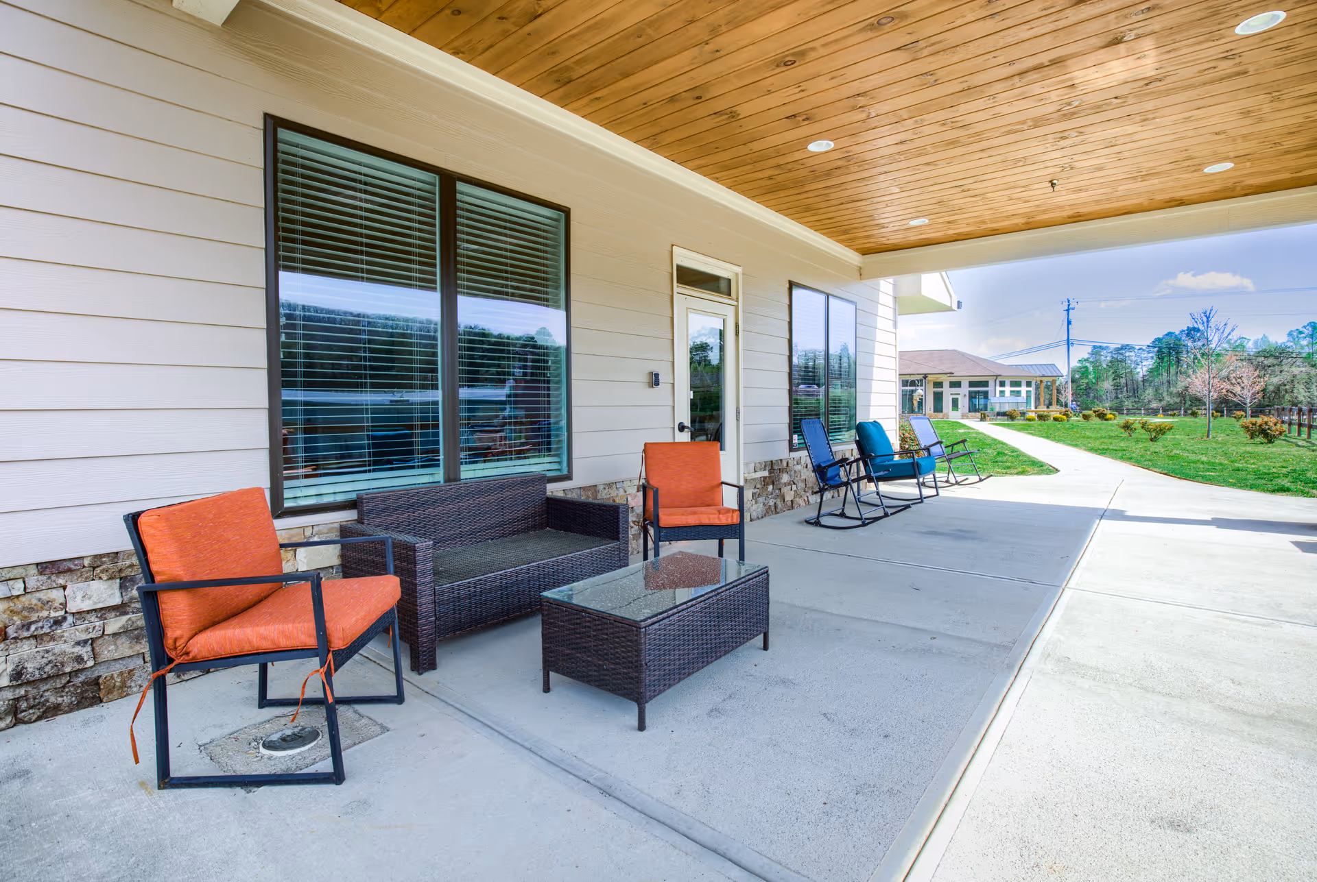 Outdoor covered patio area with wicker furniture including a loveseat, glass-top coffee table, and chairs with orange and blue cushions. The patio overlooks a green lawn and another building in the distance under a clear sky.