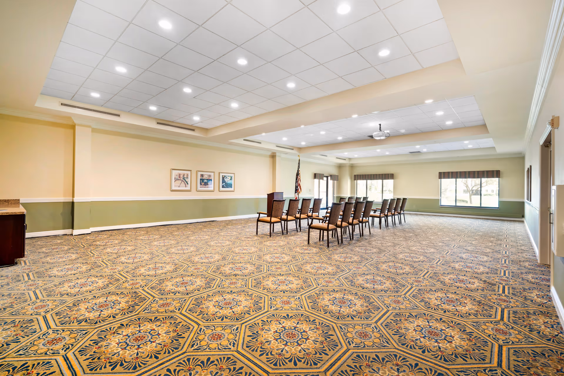 A spacious multipurpose meeting room with patterned carpet, rows of chairs facing a podium and large windows letting in natural light.
