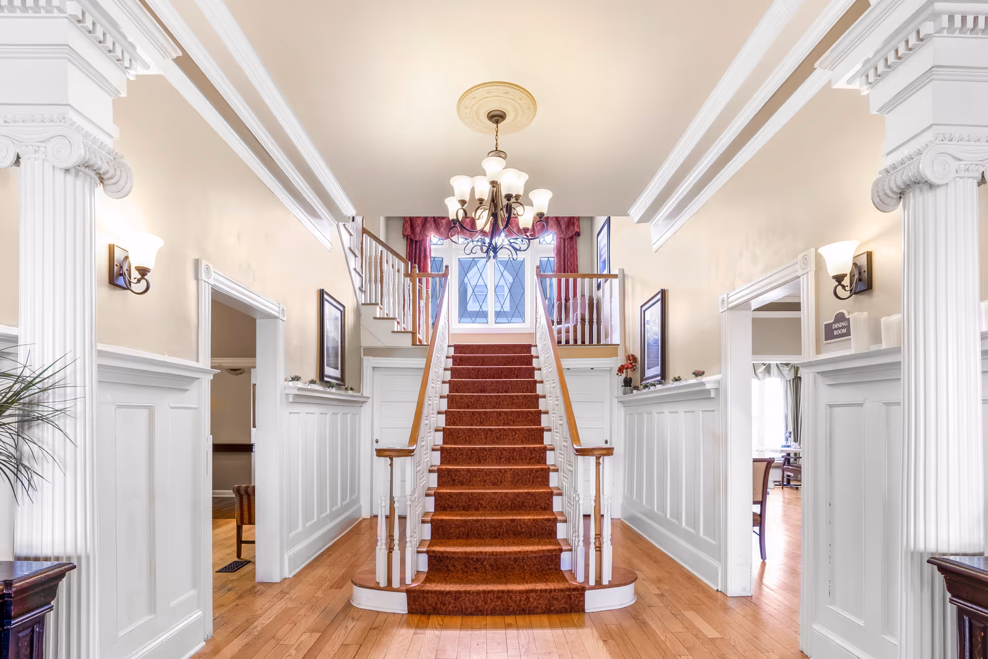 Grand foyer with a central red-carpeted staircase, white paneled walls, chandelier, and adjoining rooms.