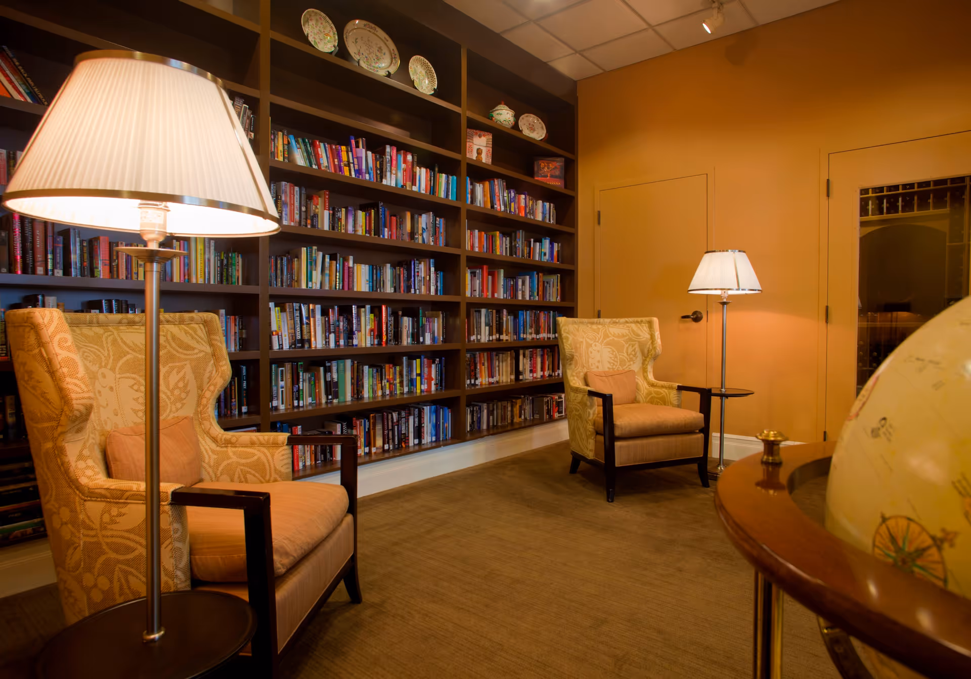 A cozy reading room with two patterned armchairs, each accompanied by a floor lamp. Behind the chairs is a large bookshelf filled with books and decorative plates on the top shelf. The walls are painted a warm yellow, and a globe on a wooden stand is partially visible in the foreground.