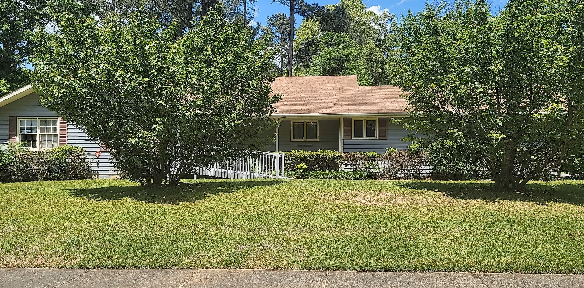 Front exterior view of a single-story residential building with a brown shingle roof and light blue siding, partially obscured by two large leafy trees and surrounded by green grass and shrubs under a blue sky with some clouds.