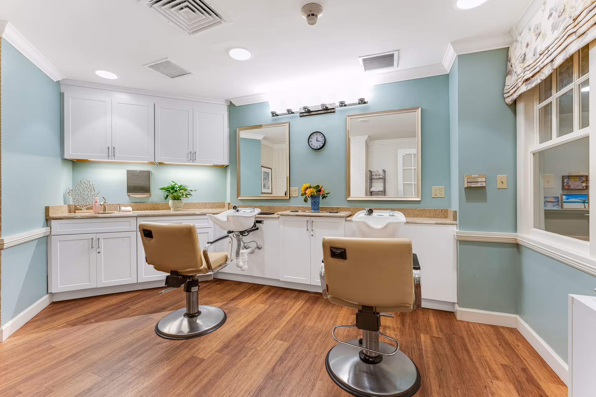 Interior view of a salon area in a senior living facility with two beige salon chairs in front of two wash basins and mirrors. The room has light blue walls, white cabinets, a wooden floor, and a window with a patterned valance.
