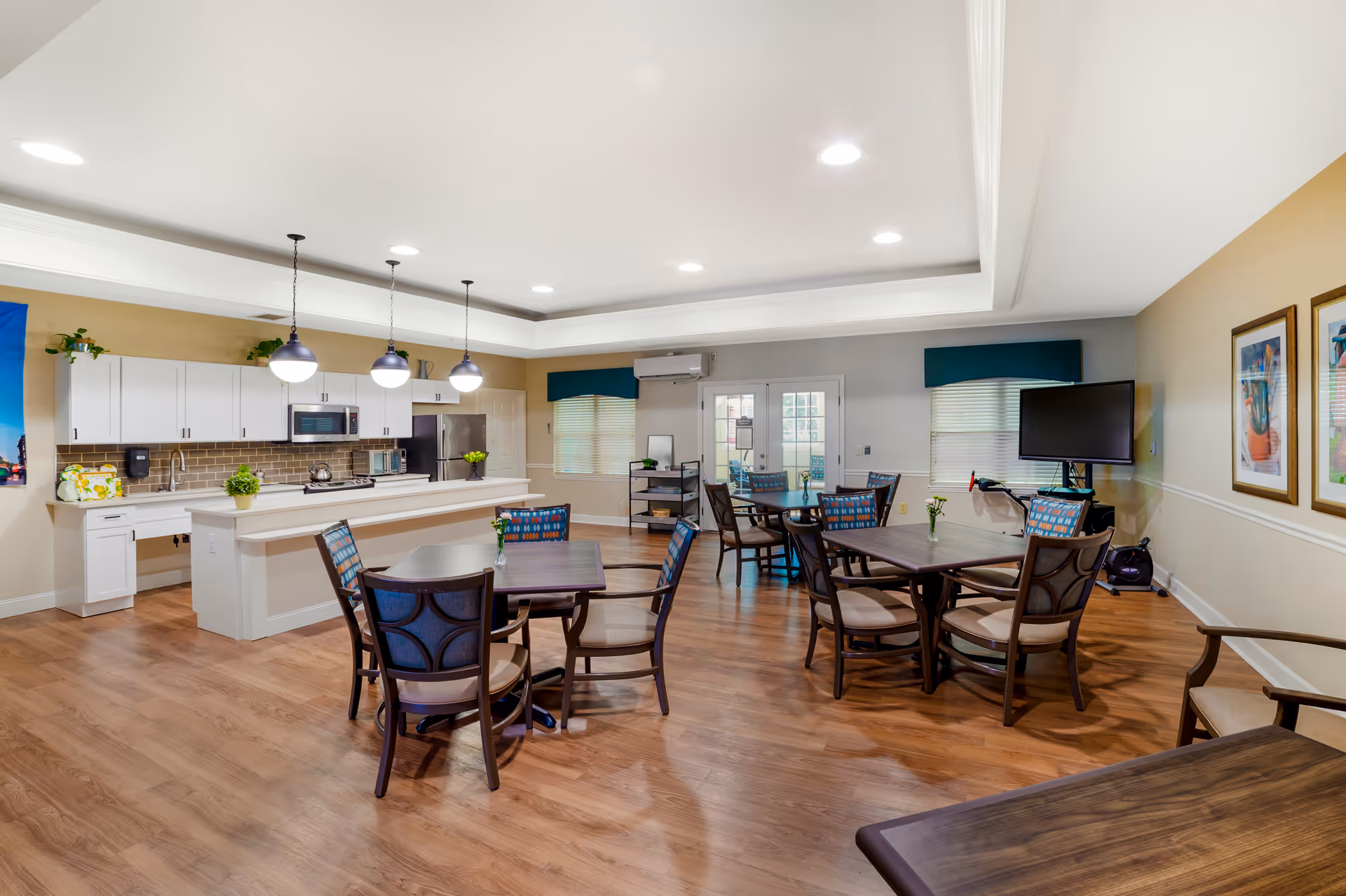 A spacious, well-lit common dining area with wooden floors, multiple tables and chairs, a kitchen area with white cabinets, a refrigerator, microwave, and pendant lights hanging over a kitchen island. The room has beige and gray walls, windows with blinds, framed artwork on the walls, and a flat-screen TV mounted on one side.