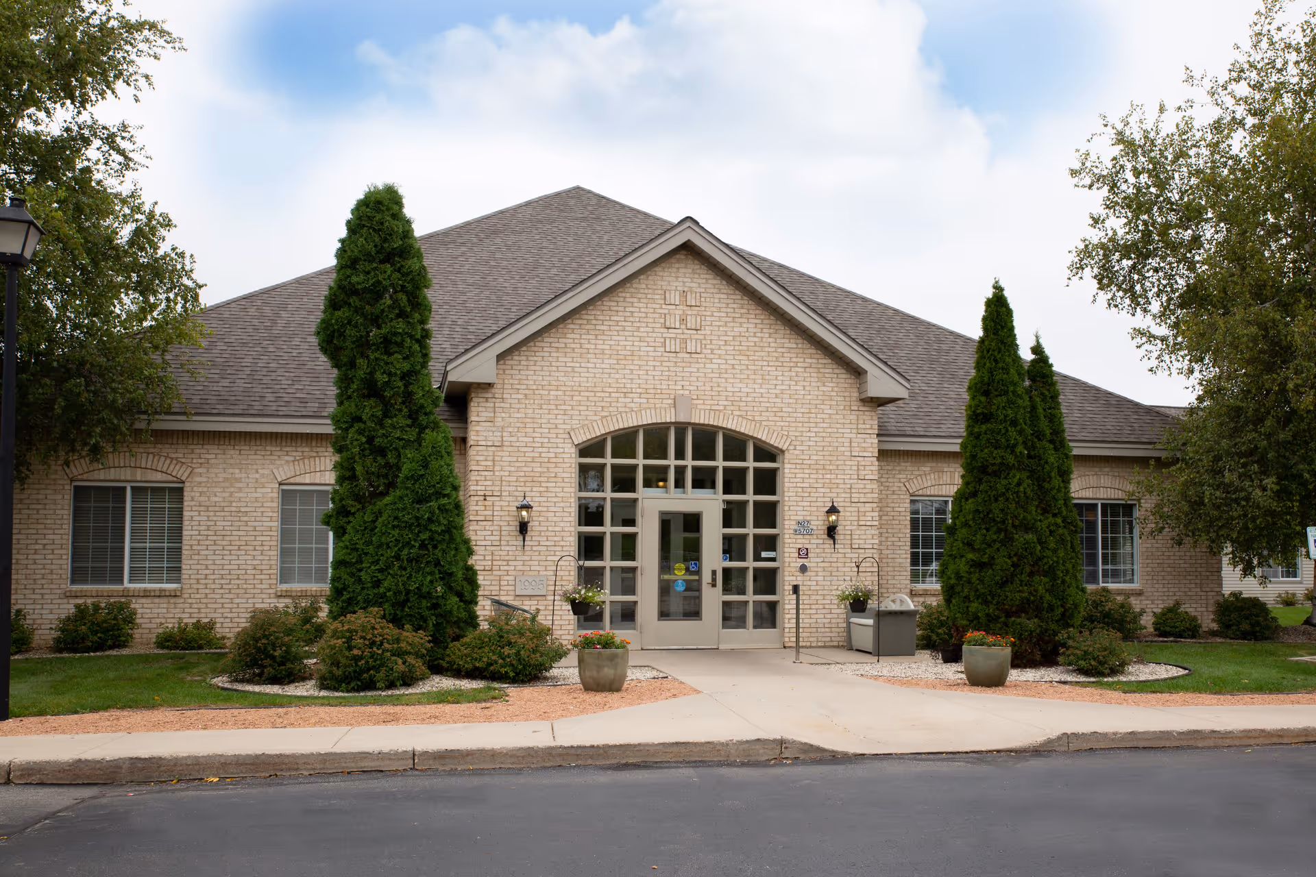 Front exterior view of a single-story brick building with a peaked roof, large central entrance with glass doors and windows, flanked by tall evergreen trees and landscaped bushes. The sky is partly cloudy.