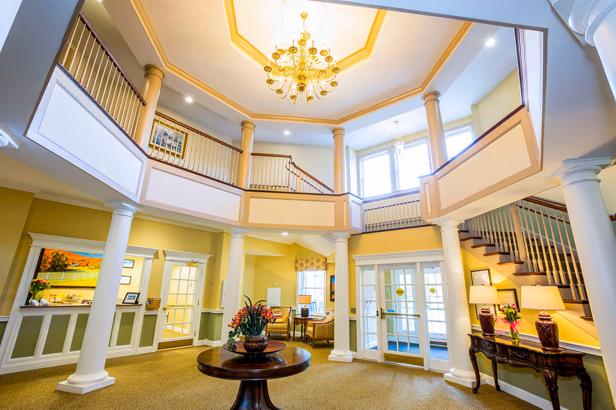 Bright and spacious senior living facility lobby with a round wooden table holding a floral arrangement in the center. The room features tall white columns, a staircase with wooden handrails, large windows letting in natural light, and a golden chandelier hanging from a decorative ceiling. There are seating areas with chairs and lamps, and double glass doors leading outside.