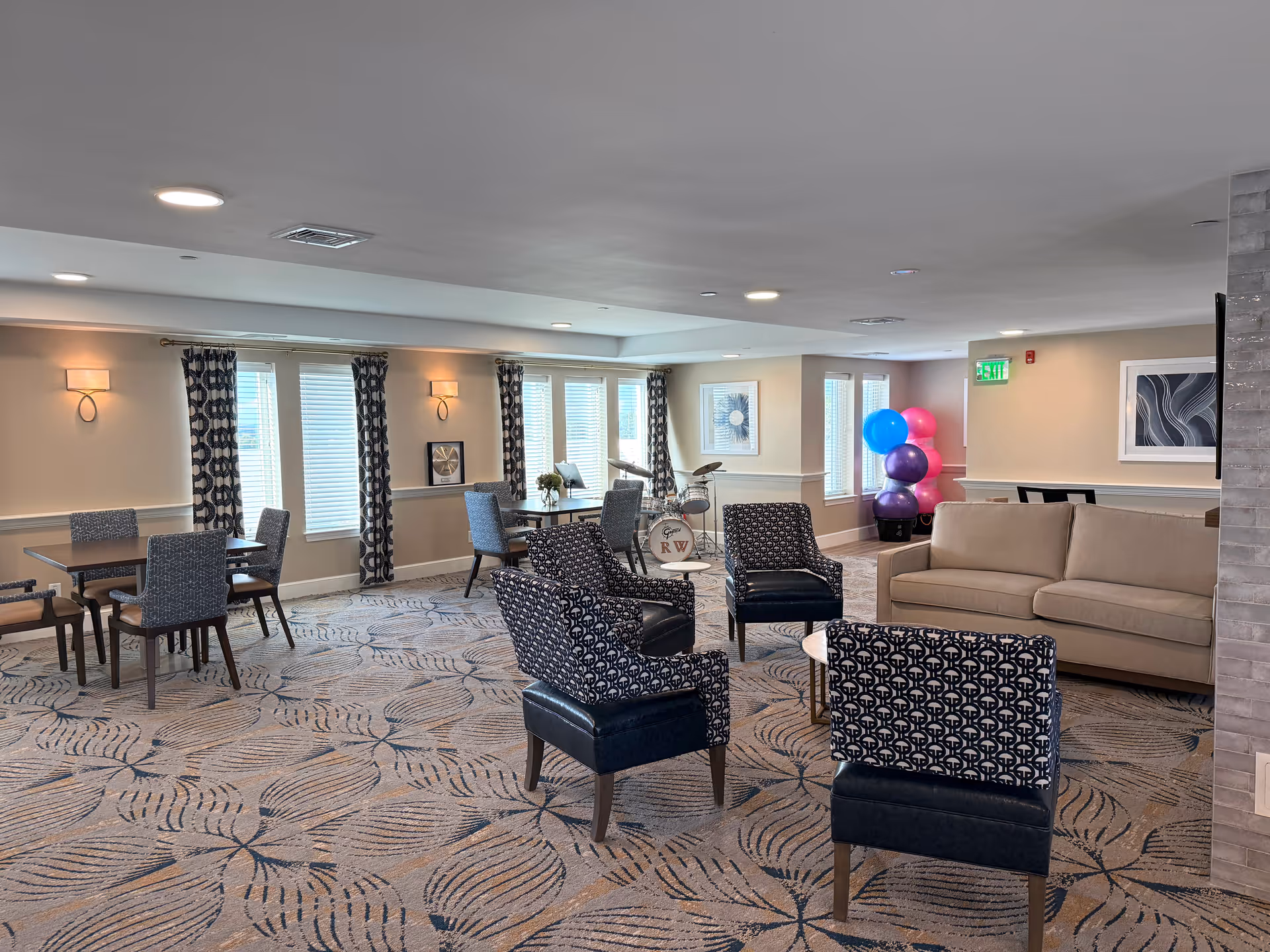 Community lounge with multiple seating areas, tables, a sofa, patterned carpet, and windows with curtains.