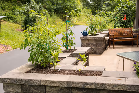 Outdoor patio area with raised garden beds containing various plants, a wooden bench, a glass-top table, and a paved walking path surrounded by greenery.