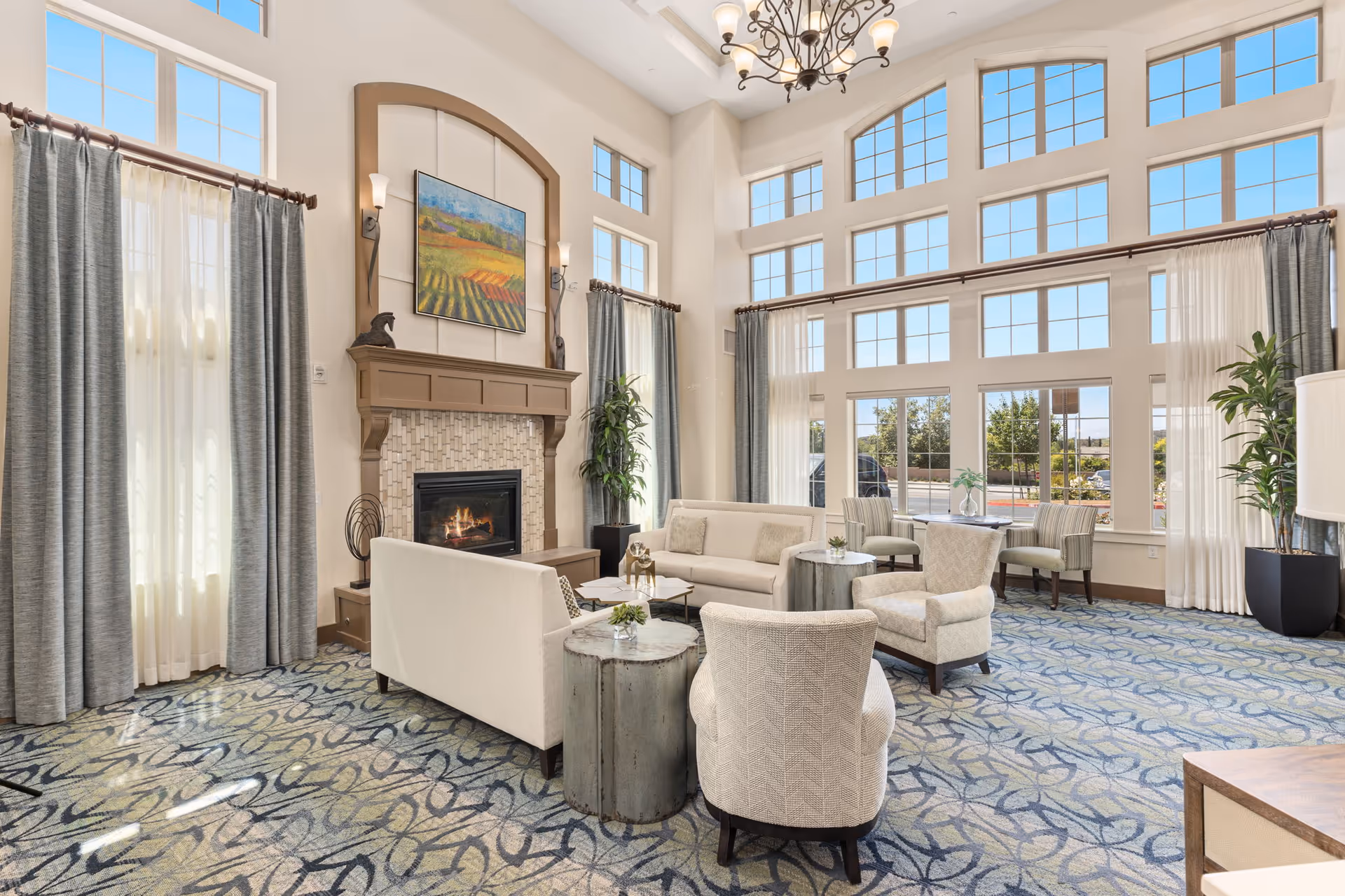 Sunlit common lounge with tall grid windows, upholstered seating arranged around a fireplace and a chandelier.