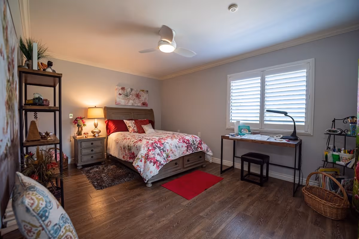 Well-decorated bedroom with a wooden bed dressed in floral bedding, nightstand and lamp, a desk under a shuttered window, shelving and hardwood floors.