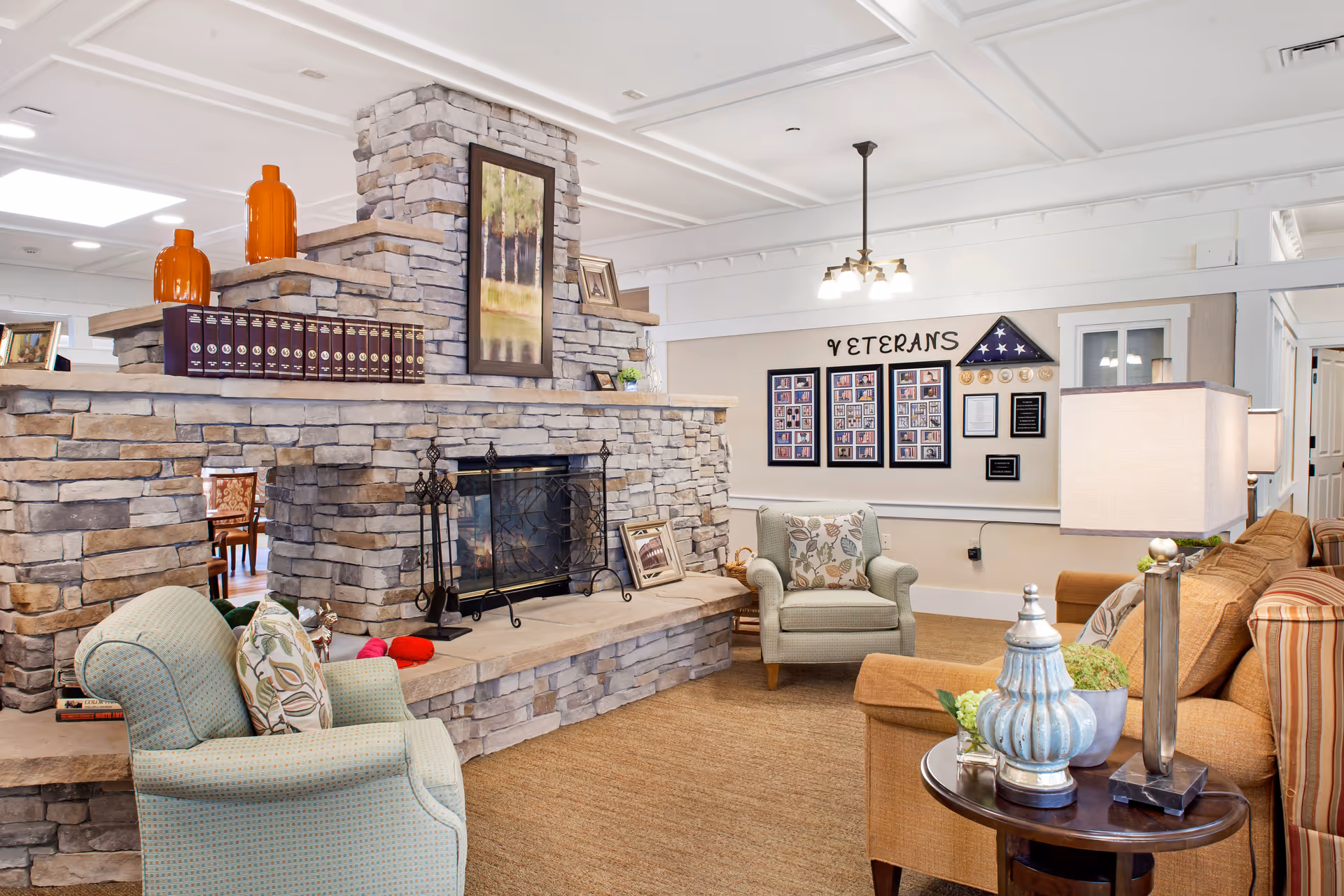 A cozy living room area in a senior living facility featuring a stone fireplace with decorative items on the mantel, two upholstered armchairs with patterned cushions, a brown sofa, a side table with a lamp and decorative pieces, and a wall display honoring veterans with framed photos and plaques.