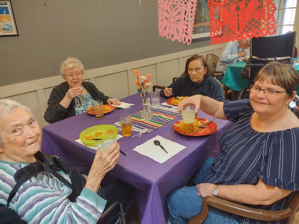 Four elderly women sitting around a table covered with a purple tablecloth, enjoying a meal together. The table has colorful plates with food, glasses of drinks, a vase with flowers, and decorative paper banners hanging above. The setting appears to be a communal dining area in a senior living facility.
