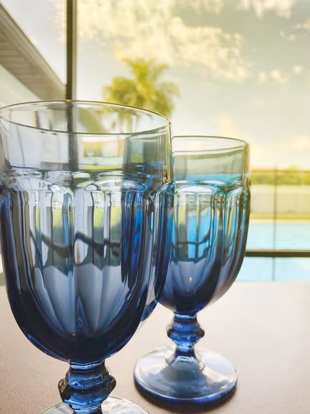 Two blue tinted glass goblets filled with water placed on a table with a blurred background showing a pool, a palm tree, and a partly cloudy sky.