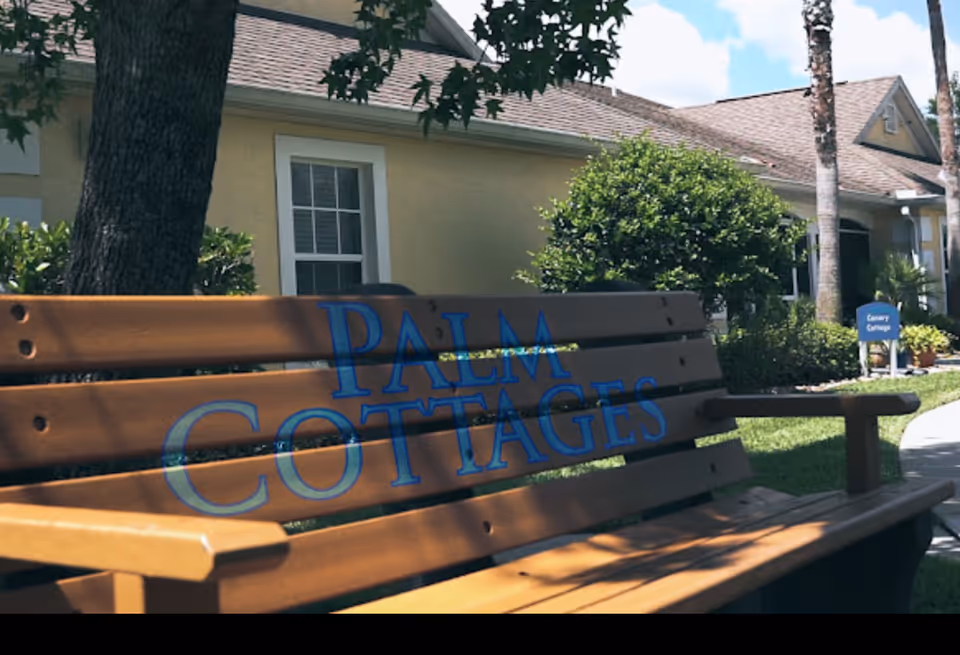 A wooden bench with the words 'Palm Cottages' painted in blue is placed outdoors near a tree. In the background, there is a yellow building with white-trimmed windows, green bushes, palm trees, and a blue sign that reads 'Canary Cottage'. The sky is partly cloudy.