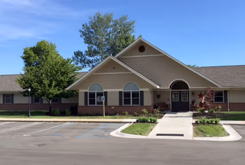 Front entrance of a single-story residential building with a gabled roof, walkway, parking spaces, and landscaping under a clear sky.