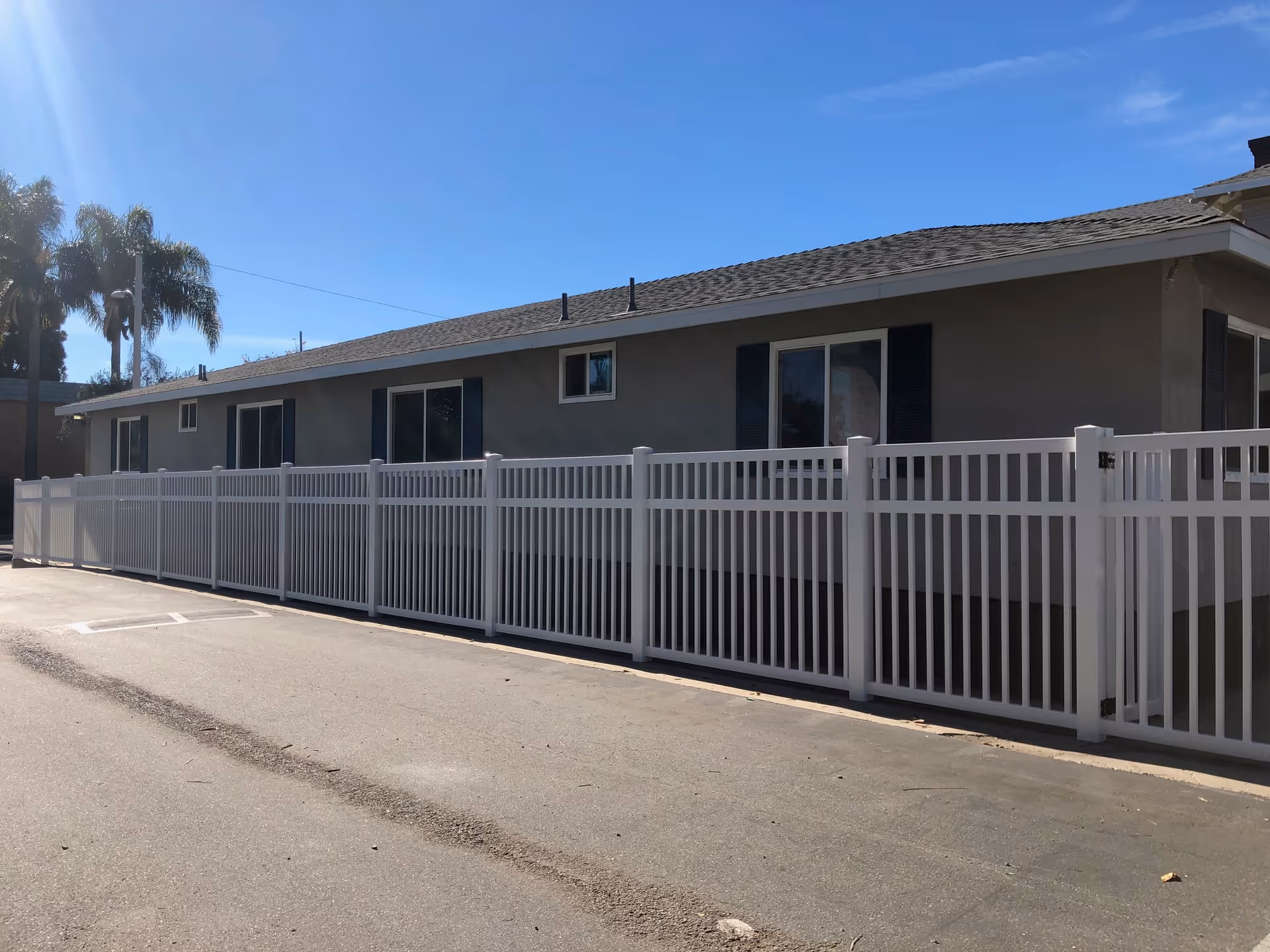 Single-story building with a white vinyl fence along a driveway under a clear blue sky.