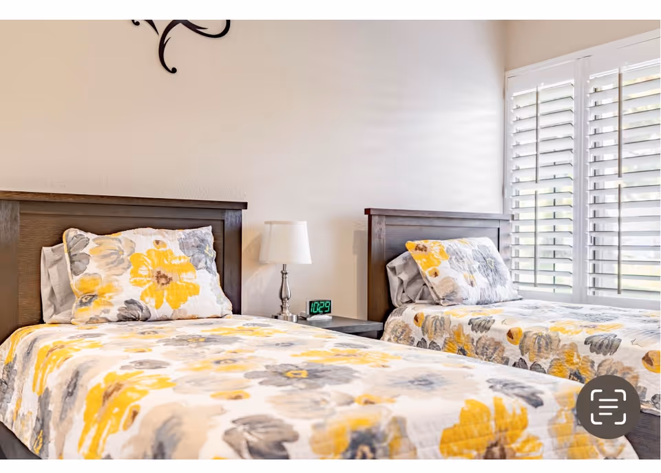 A bright bedroom with two twin beds featuring matching floral bedspreads in yellow, gray, and white. Between the beds is a small nightstand with a silver lamp and a digital clock showing 10:29. The room has white walls and a window with white plantation shutters allowing natural light to enter.