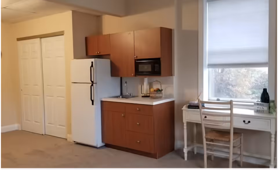 A small kitchenette with a refrigerator, microwave, sink and wood cabinets next to a desk and chair by a window.