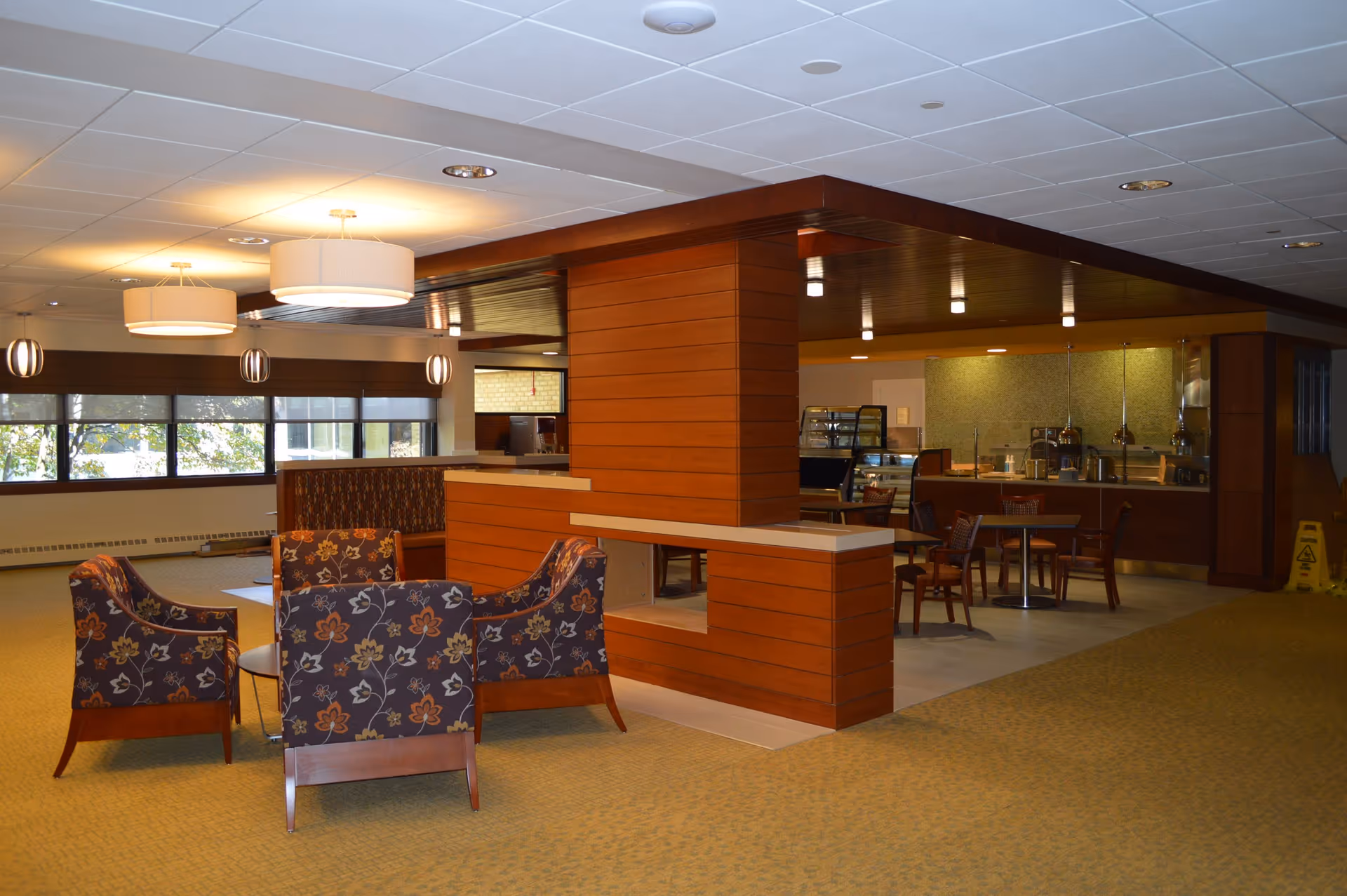 Interior view of a senior living facility lounge area with floral patterned armchairs arranged around a small round table. The space features warm wood paneling, large windows with roller shades, modern ceiling lights, and a dining area with tables and chairs in the background near a food service counter.