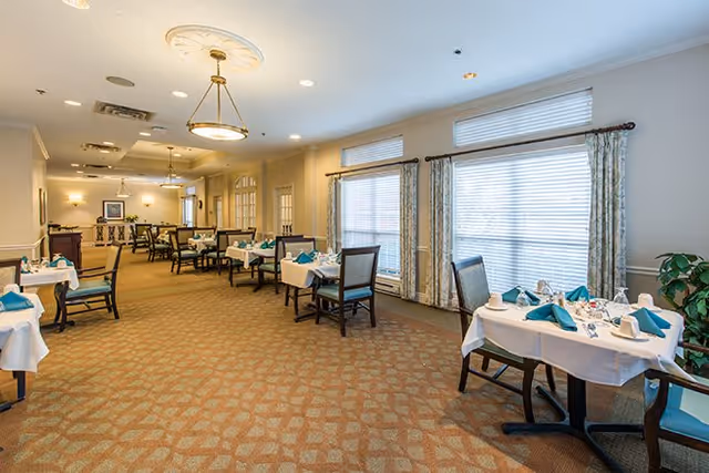 A spacious dining room with multiple tables set with white tablecloths, teal napkins, and dining ware. The room has large windows with blinds and patterned curtains, beige walls, and carpeted flooring. There are several chairs around each table and ceiling light fixtures providing warm lighting.