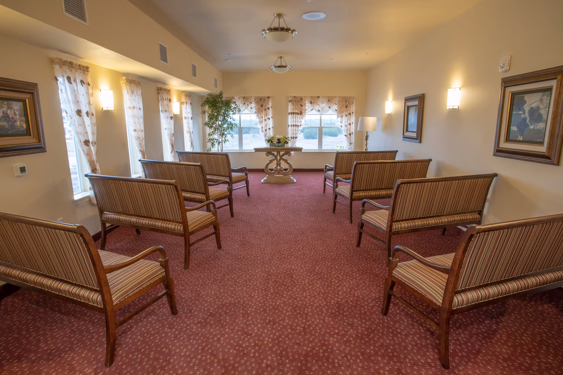 Carpeted communal seating room with rows of striped wooden benches facing a decorative table beneath a window.