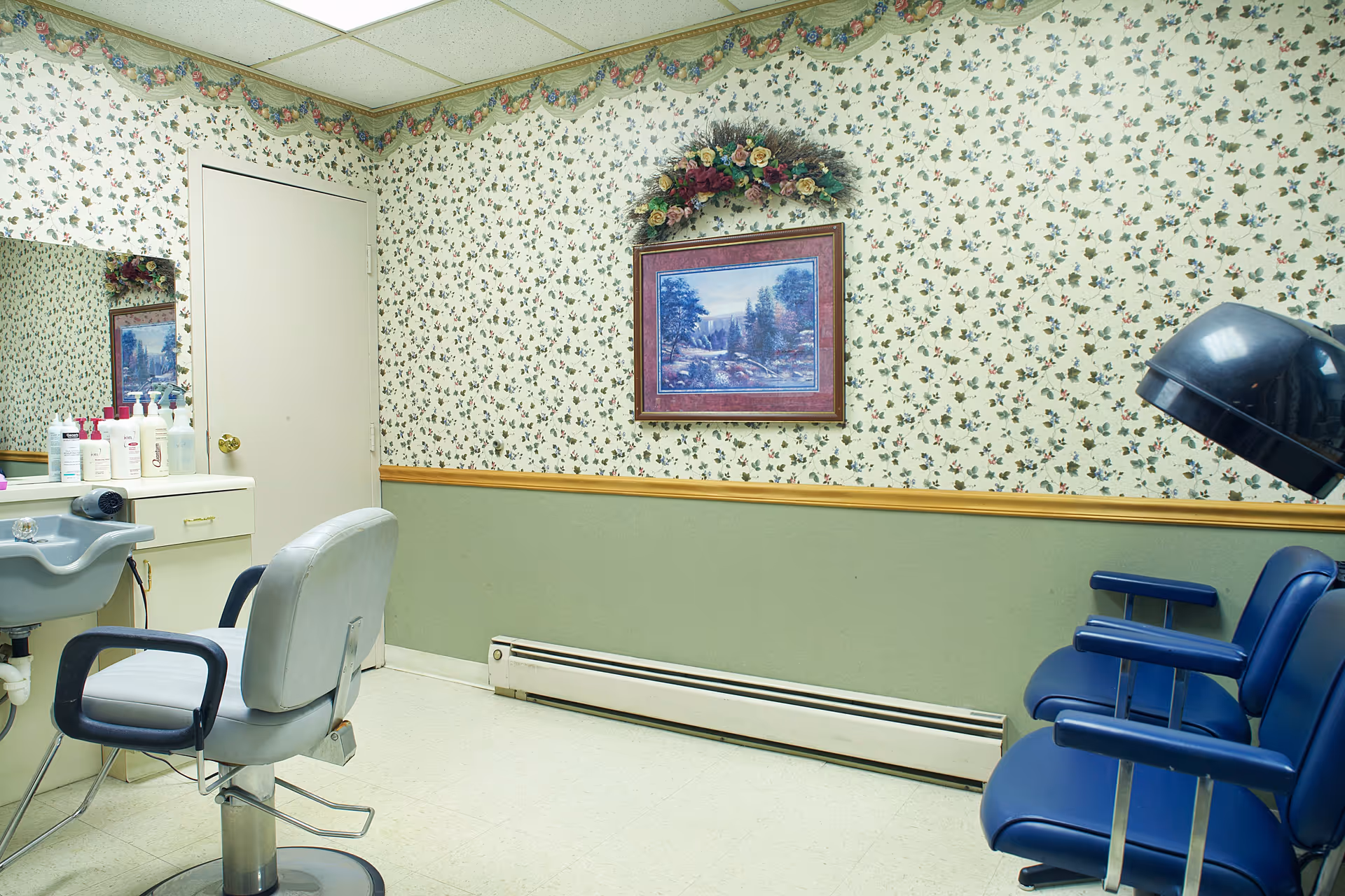 Interior of a small salon room with floral wallpaper and green lower wall paneling. There is a salon chair in front of a sink and counter with various hair care products. On the right side, there are two blue salon chairs with a black hair dryer hood. A framed landscape painting with a floral wreath above it hangs on the wall.