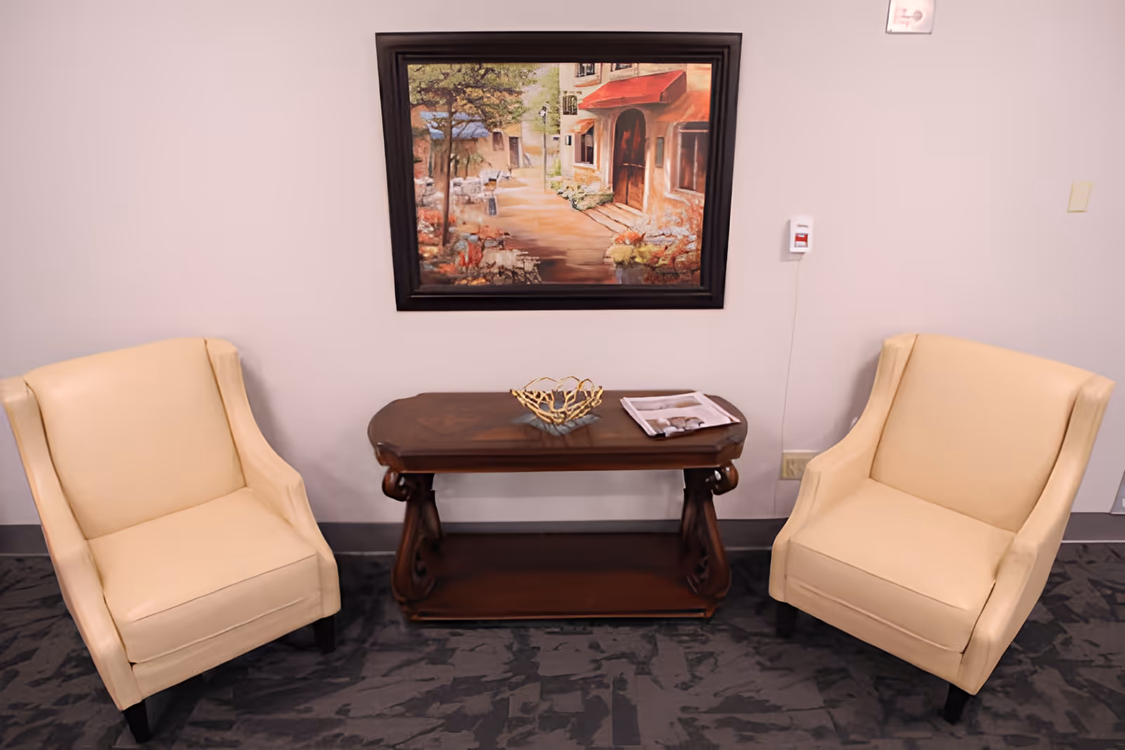 Two beige armchairs flank a dark wooden console table topped with a decorative bowl and magazine beneath a framed painting on a pale wall.