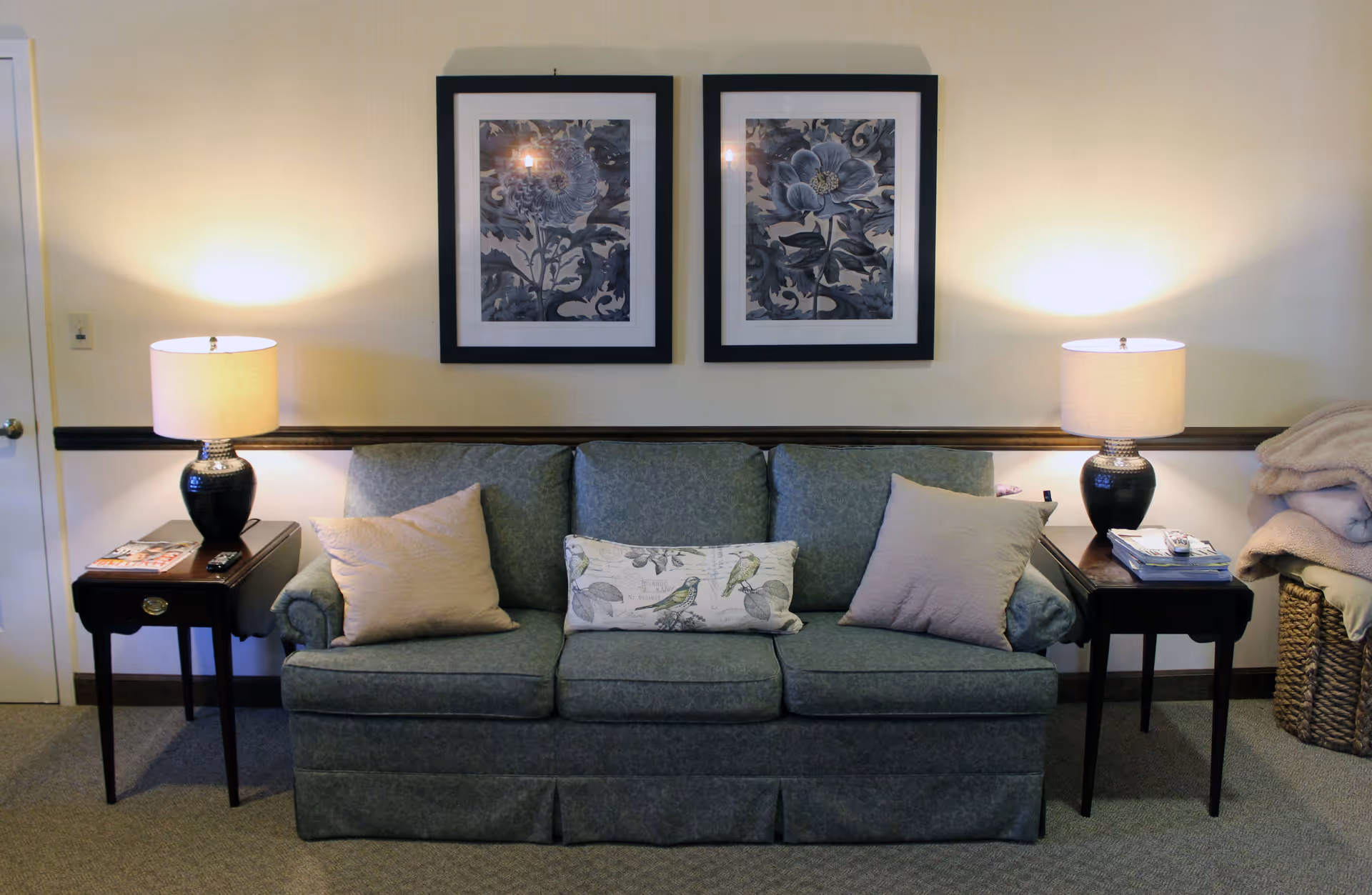 Green upholstered sofa with throw pillows between two side tables and lamps, and two framed floral prints on the wall above.