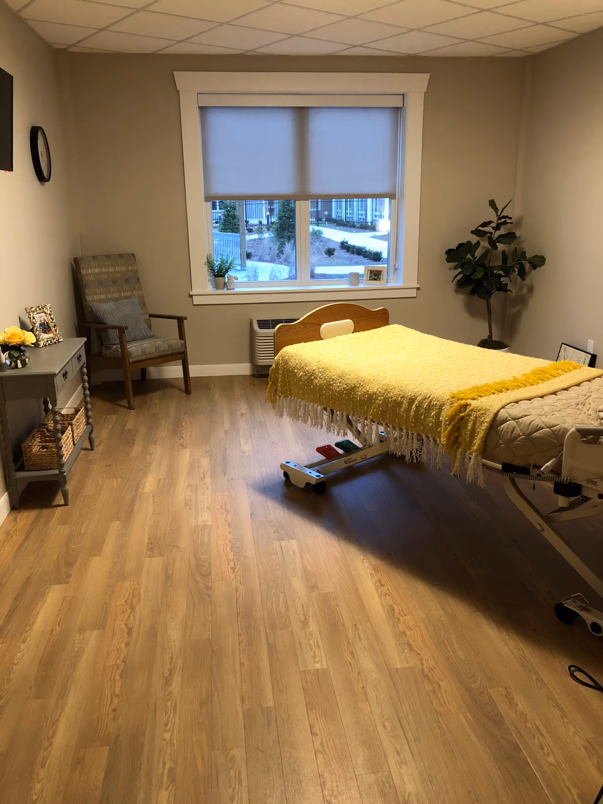 A well-lit room with a medical bed covered with a yellow blanket, a wooden chair with a cushion, a small table with decorative items including a flower vase and framed photos, a large window with a roller shade partially down, and a potted plant in the corner. The floor is wooden, and the walls are painted beige.