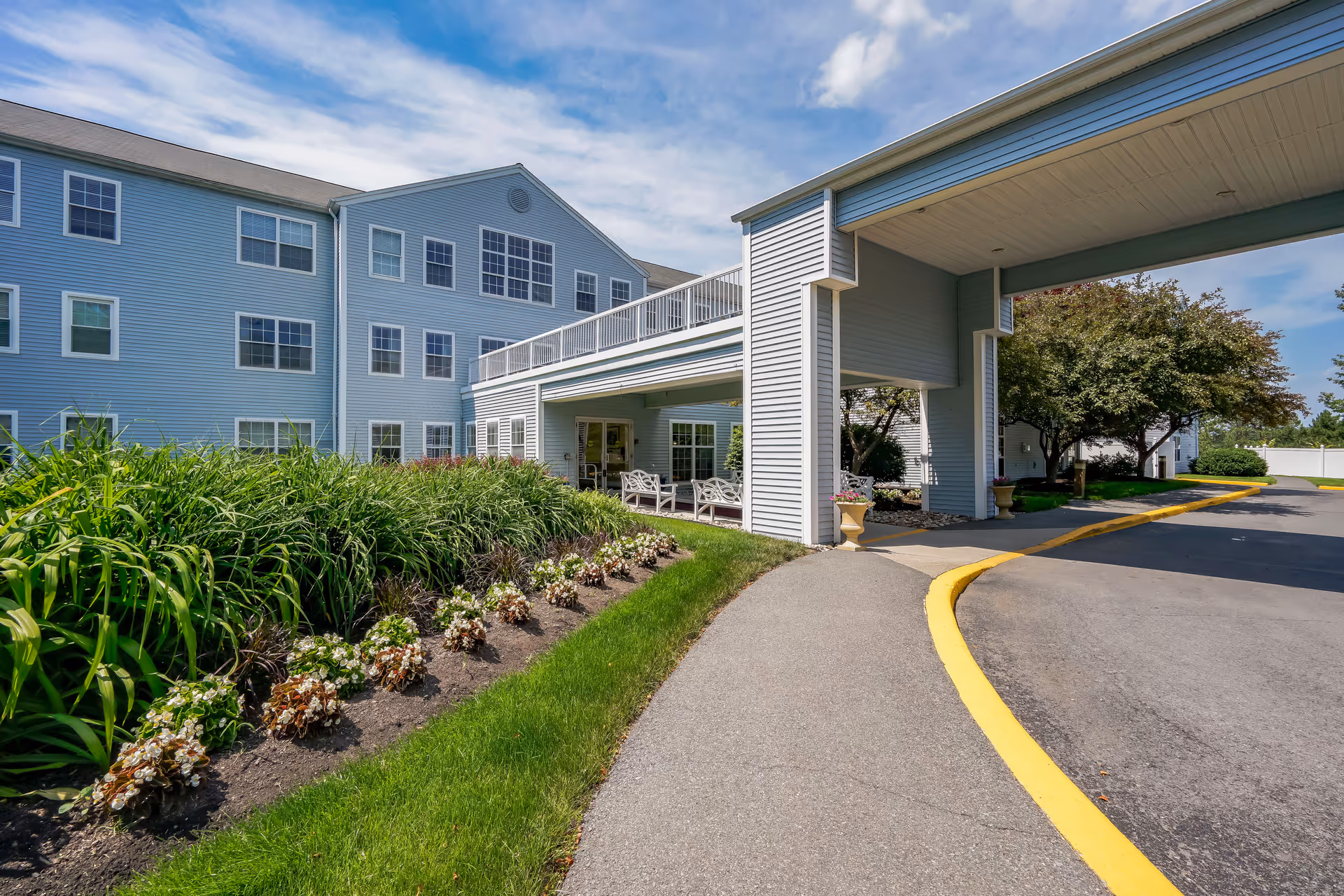 Exterior view of a senior living community building with light blue siding, multiple windows, and a covered entrance driveway. There are green shrubs and flower beds along the sidewalk leading to the entrance, with a clear blue sky and some clouds overhead.