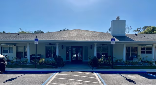 Front entrance of a single-story senior living building with a covered porch, double doors, and two handicap parking signs and spaces in front.