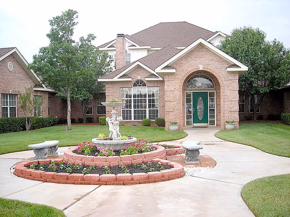 Front exterior view of a brick building with a green door, large windows, and a peaked roof. In front of the building is a circular flower bed with a decorative fountain featuring cherubs, surrounded by blooming flowers and two stone benches. The area is landscaped with green grass, trees, and shrubs.