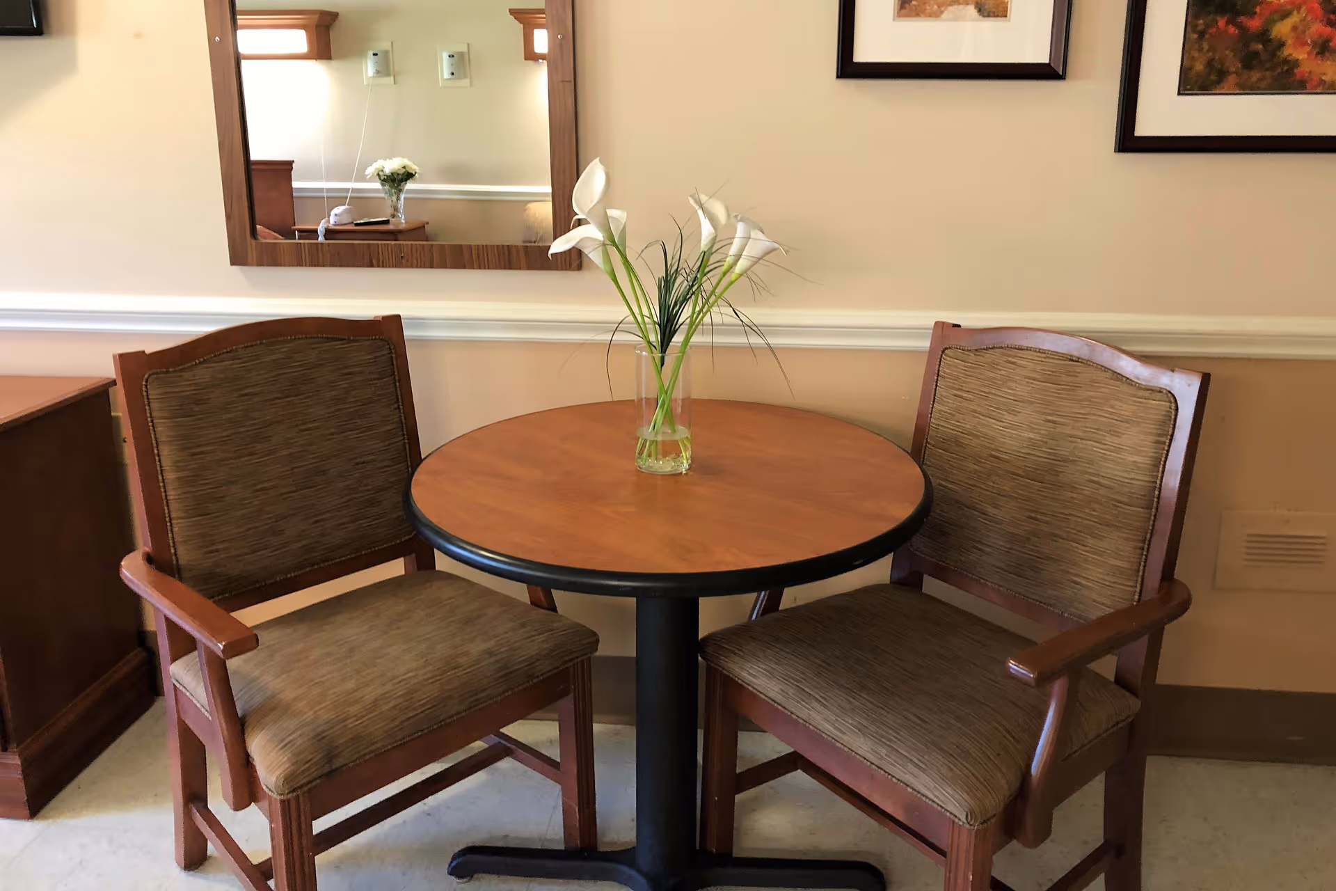 A small round wooden table with a glass vase holding white calla lilies in the center, flanked by two wooden chairs with cushioned seats and backs. A mirror and framed pictures hang on the beige wall behind the table.